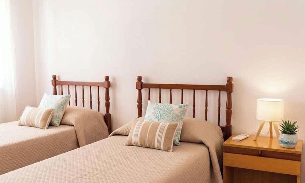 Una camera d'albergo con due letti singoli, ciascuno dotato di una testata in legno, un comodino con una lampada e un piccolo vaso con una pianta.