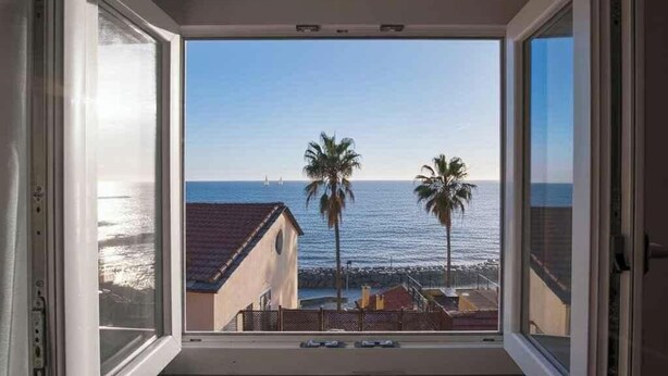 A window with a view of the ocean, palm trees, and a building with a red roof. - Holiday apartment with air conditioning (Imperia)