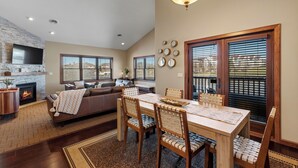 A modern living room with a fireplace, a dining area with wooden chairs, a ceiling fan, and a large window with blinds. - Riverfront Condo Steps to Downtown & Lake Michigan (Sheboygan)