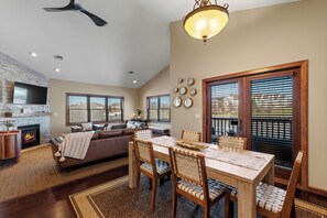 Ein modernes Wohnzimmer mit Kamin, Essbereich mit Holzstühlen, Deckenventilator und großem Fenster mit Jalousien.