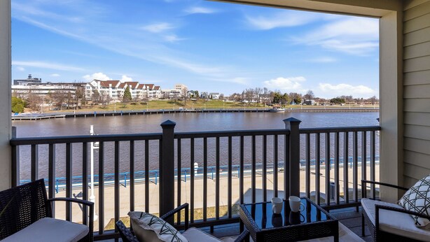 A balcony with outdoor furniture overlooking a river, a dock, and buildings in the background. - Riverfront Condo Steps to Downtown & Lake Michigan (Sheboygan)