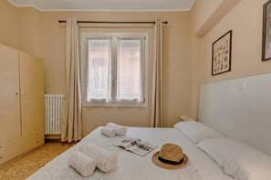 Ein Hotelzimmer mit Bett, Kleiderschrank, Heizkörper, einem Fenster mit Vorhängen und einem Hut auf dem Bett.