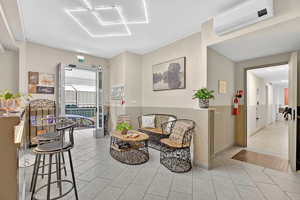 Eine Lobby mit einer Rezeption, zwei Barhocker, einer Couch und einem Couchtisch. An den Wänden befinden sich gerahmte Bilder, ein Blumentopf und ein Feuerlöscher.