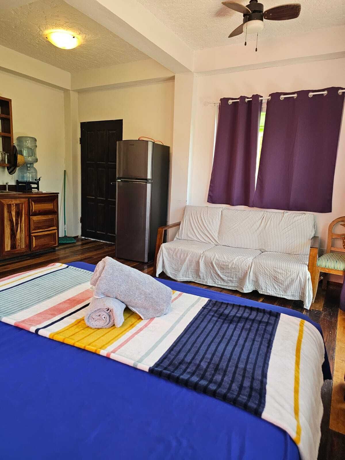 Une chambre à coucher comprenant un lit, un ventilateur de plafond, un réfrigérateur, un distributeur d’eau et une fenêtre avec des rideaux.