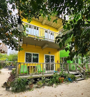Un edificio giallo di due piani con un balcone, circondato da vegetazione e piante in vaso.