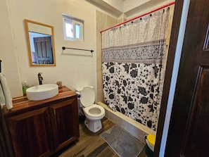 Un bagno con un mobile vanitario in legno, un lavandino bianco, un water, una tenda da doccia con motivo floreale e una piccola finestra sopra il water.