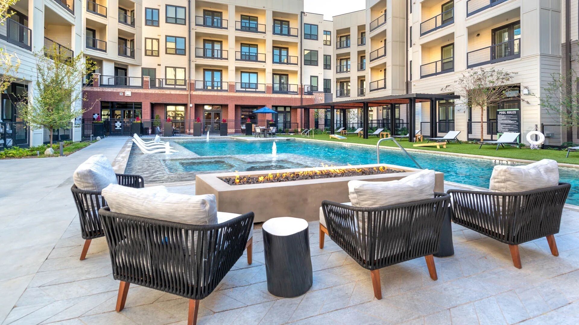 A pool area with lounge chairs and a fire pit, surrounded by a multi-story building with balconies.