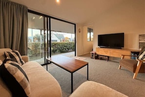 Uma sala de estar com sofá, mesa de centro, televisão e porta de vidro deslizante que dá para uma varanda.