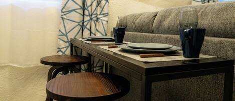 A dining area with a table set for two, two stools, a sofa, and a patterned curtain.