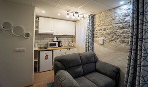 Eine kompakte Küchenzeile mit Mikrowelle, Wasserkocher und Kaffeemaschine, einem kleinen Kühlschrank sowie einem grauen Sofa in einem Raum mit Steinwänden und einem Vorhang.
