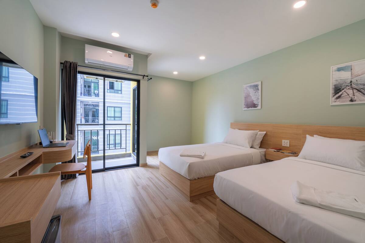 A hotel room with two beds, a desk with a laptop, a chair, and a large window with a view of buildings.