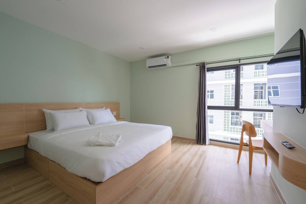 A hotel room with a bed, a desk, a chair, and a large window with a view of buildings.