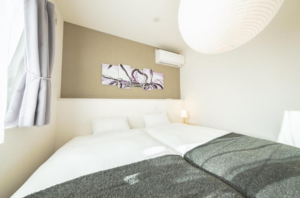 A hotel room with a bed, a wall-mounted air conditioner, a painting, and a lamp.