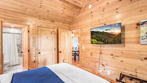 Ein Zimmer mit Holzwandverkleidung, einem Bett, einem an der Wand befestigten Fernseher und einem durch eine offene Tür einsehbaren Badezimmer.