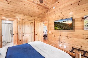 A wooden-paneled room with a bed, a television mounted on the wall, and a bathroom visible through an open door. - Cozy Cabin with Great Views • Hot Tub • Wi-Fi (Murphy)
