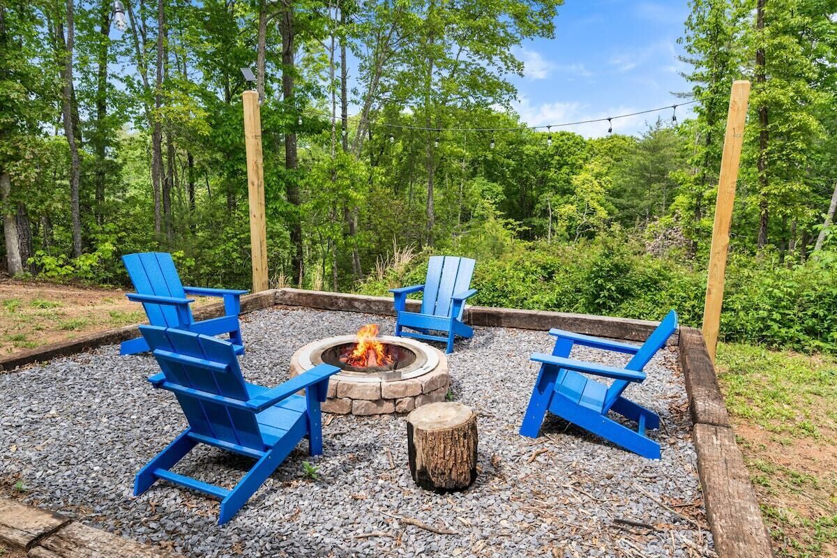 A fire pit with four blue Adirondack chairs arranged around it, set on a gravel surface with a wooden border.