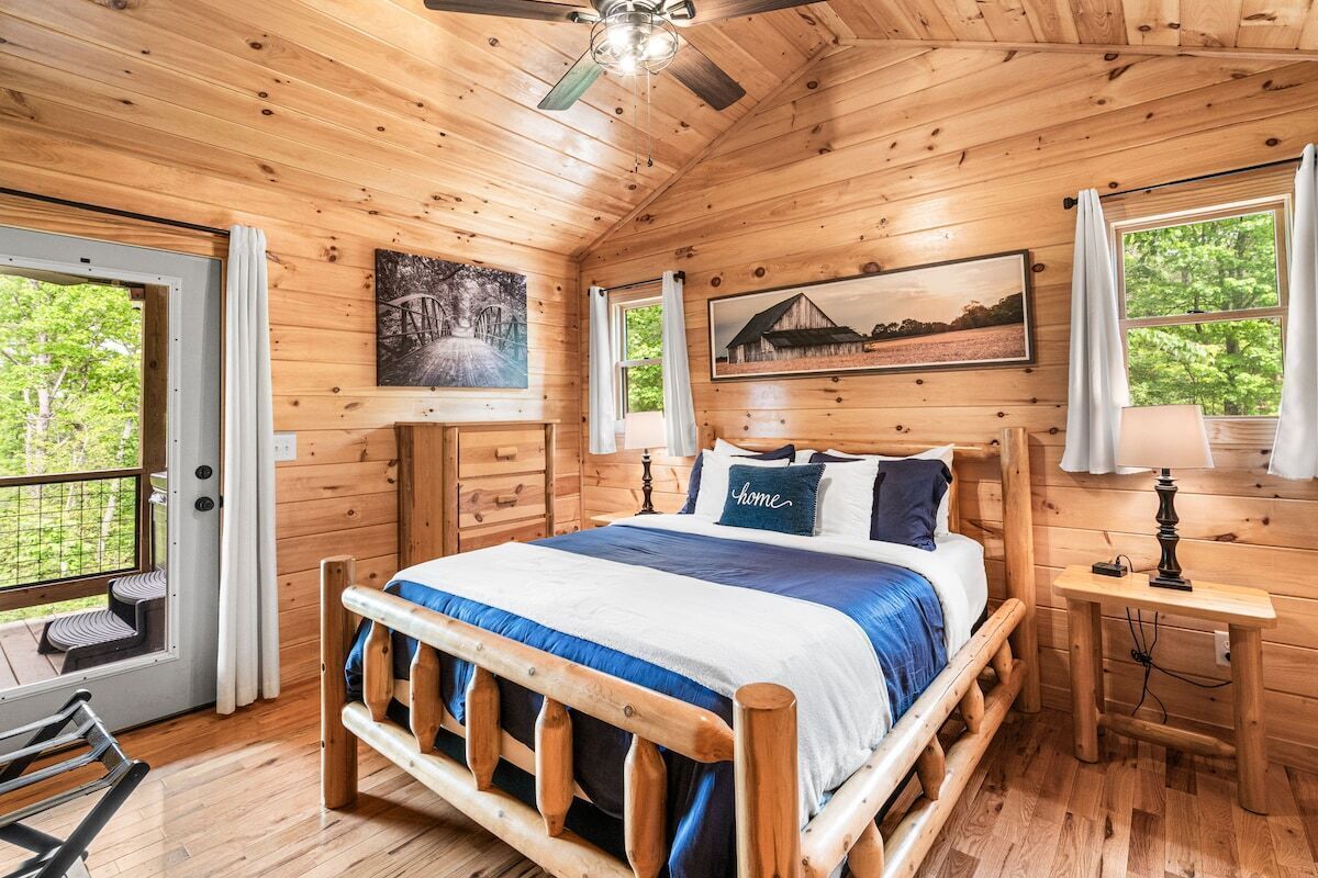 A wooden cabin bedroom with a bed, two windows, a ceiling fan, and artwork on the walls.