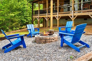 Eine Holzdiele mit Terrasse, umgeben von einem Wald. Blaue Adirondack-Stühle und eine Feuerstelle aus Stein sind vorhanden.