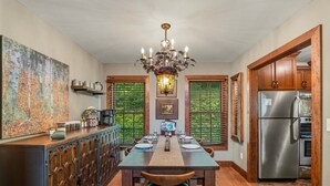 A kitchen with a dining table, chairs, a chandelier, a refrigerator, and a window with a view of greenery.