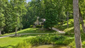 A house surrounded by a forest with a pond in the foreground.