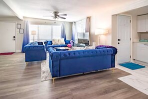 Una sala de estar con un sofá azul, un ventilador de techo y una zona de cocina con armarios.