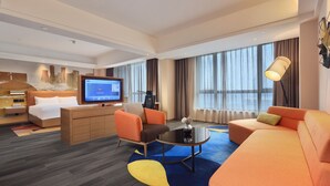Une chambre d’hôtel avec un lit, une télévision, un canapé, un fauteuil, une table basse et une grande fenêtre.