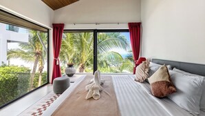 Ein modernes Hotelzimmer mit großem Fenster, einem Bett mit gestreifter Bettwäsche, einem Nachttisch und Blick auf Palmen und ein Gebäude.