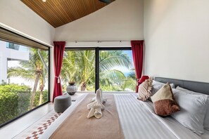 Ein modernes Hotelzimmer mit großem Fenster, einem Bett mit gestreifter Bettwäsche, einem Nachttisch und Blick auf Palmen und ein Gebäude.