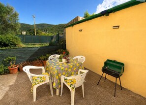 Een terras met een gele muur, een tafel met stoelen, plantenbakken en een barbecue.
