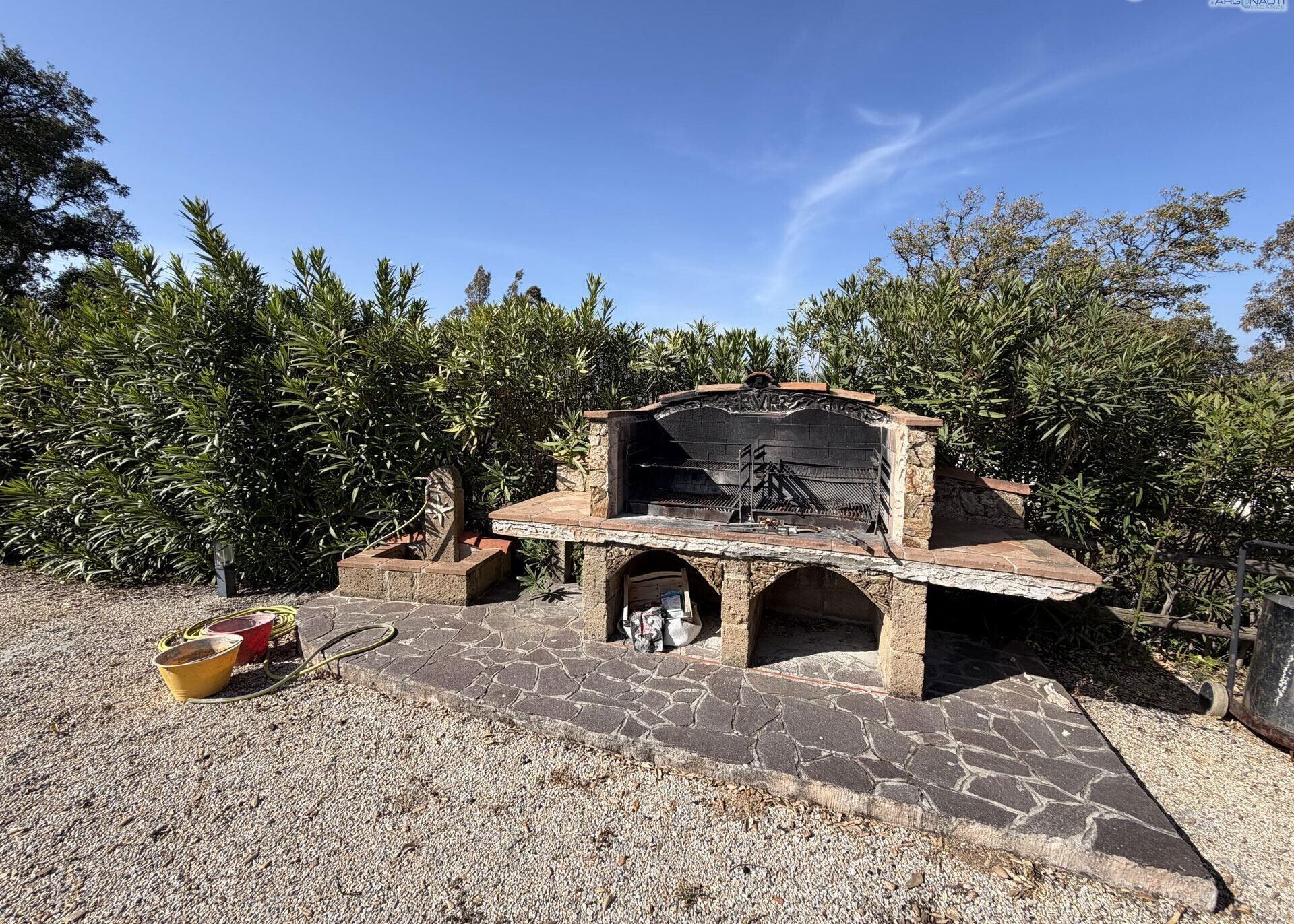 An outdoor stone barbecue with a chimney, surrounded by greenery.