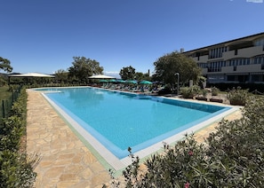 Ein rechteckiger Pool mit einer Steinterrasse, umgeben von Liegestühlen und Sonnenschirmen, mit einem Gebäude im Hintergrund.