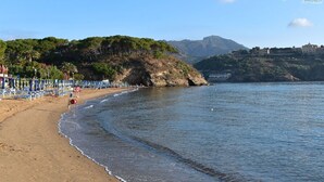 A beach with a row of sun loungers and umbrellas, a rocky outcrop, and a building in the background. - Residenza Verde Blu - Verde Blu Primo Piano (620) (Capoliveri)
