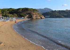 Ein Strand mit einer Reihe von Liegestühlen und Sonnenschirmen, einem Felsen und einem Gebäude im Hintergrund.