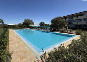 Ein rechteckiger Pool mit einer Steinterrasse, umgeben von Liegestühlen und Sonnenschirmen, mit einem Gebäude im Hintergrund.
