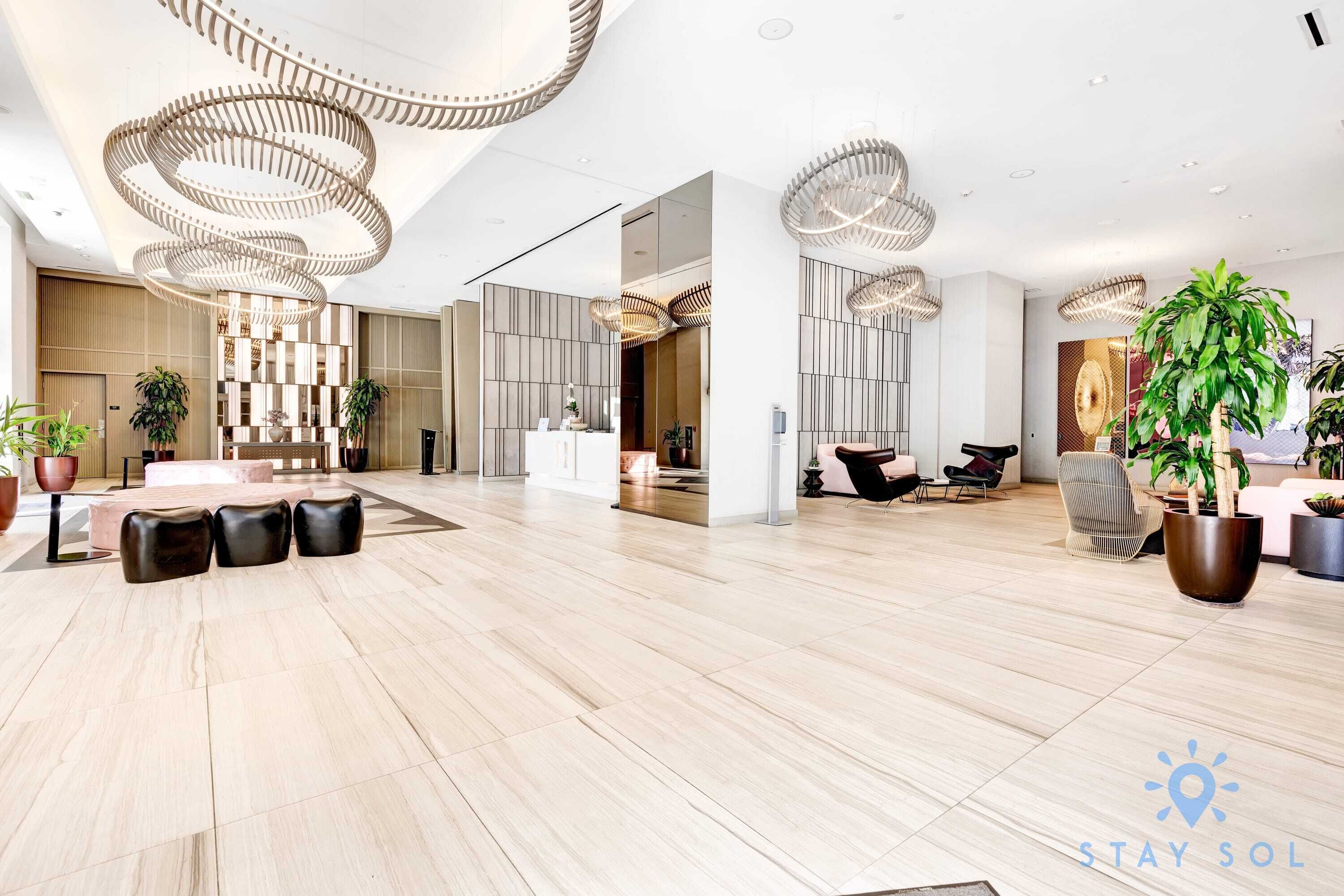 Een ruime lobby met een modern design, met een centraal zitgedeelte met een tafel en stoelen, een receptiebalie en decoratieve verlichting.