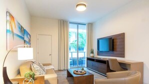 Un salon moderne avec un canapé, une table basse et un téléviseur. Il y a un balcon avec des rideaux et une vue sur des palmiers.