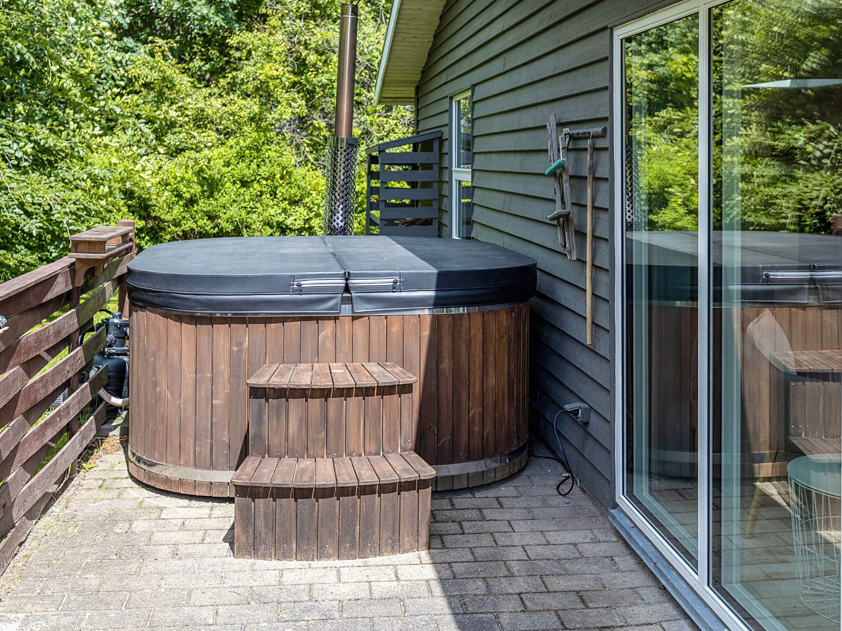 A hot tub with a wooden exterior and steps, located on a paved patio adjacent to a house with a glass door.