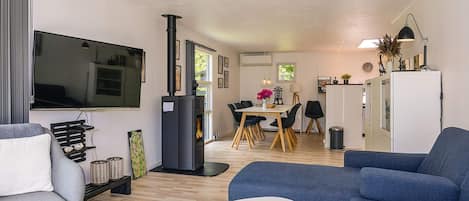 A modern living room with a flat-screen TV, a fireplace, a dining area with a table and chairs, and a kitchen area with a refrigerator.