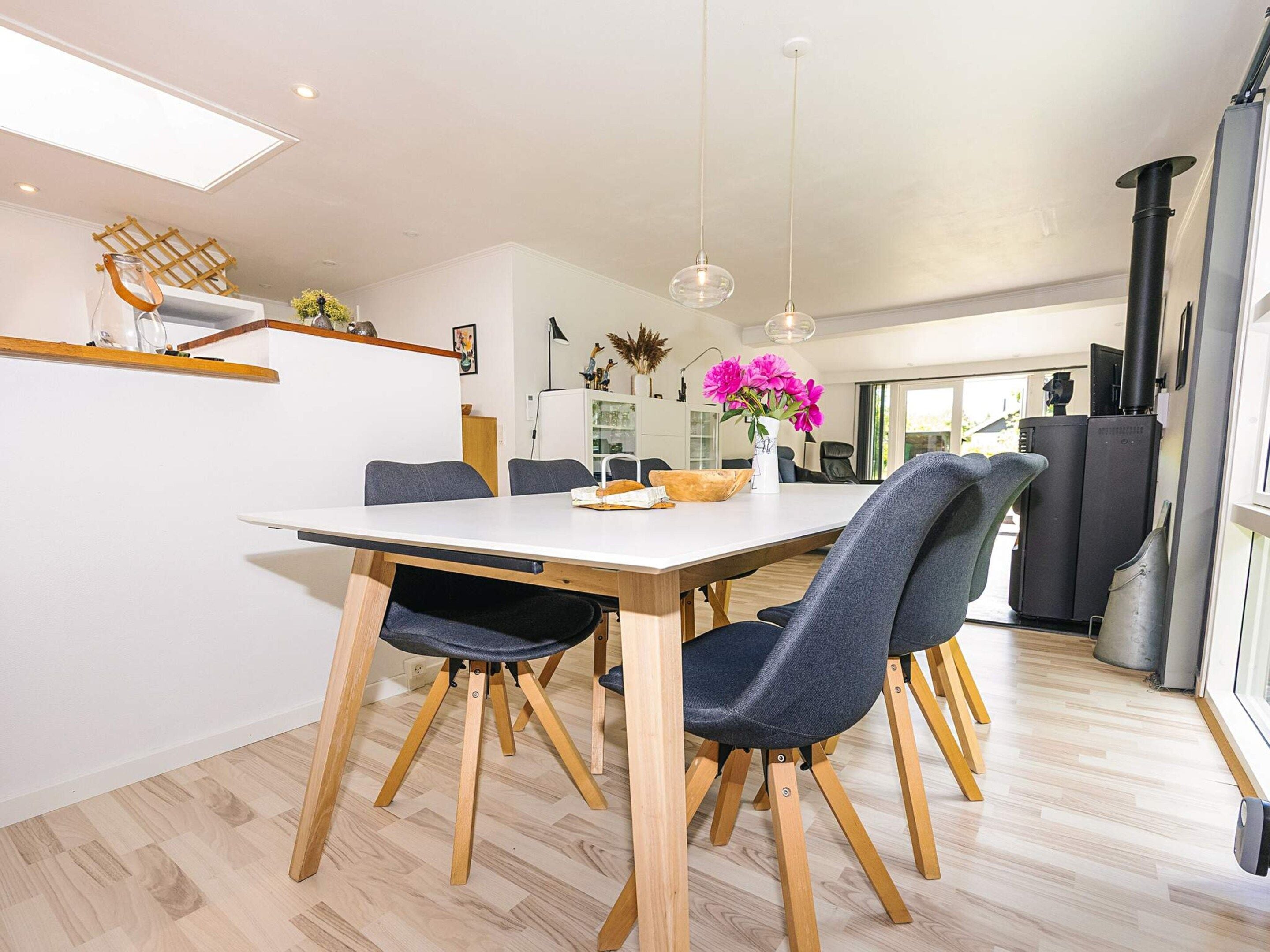 Een moderne eetruimte met een houten tafel, grijs gestoffeerde stoelen, hanglampen en een uitzicht op de keuken op de achtergrond.