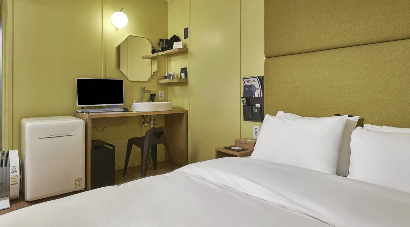 Habitación de hotel con cama, escritorio con computadora, refrigerador pequeño, espejo y repisas con artículos de tocador.