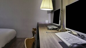 Habitación de hotel con un escritorio de madera, dos monitores, un teclado, un ratón, una lámpara, una silla y una cama.