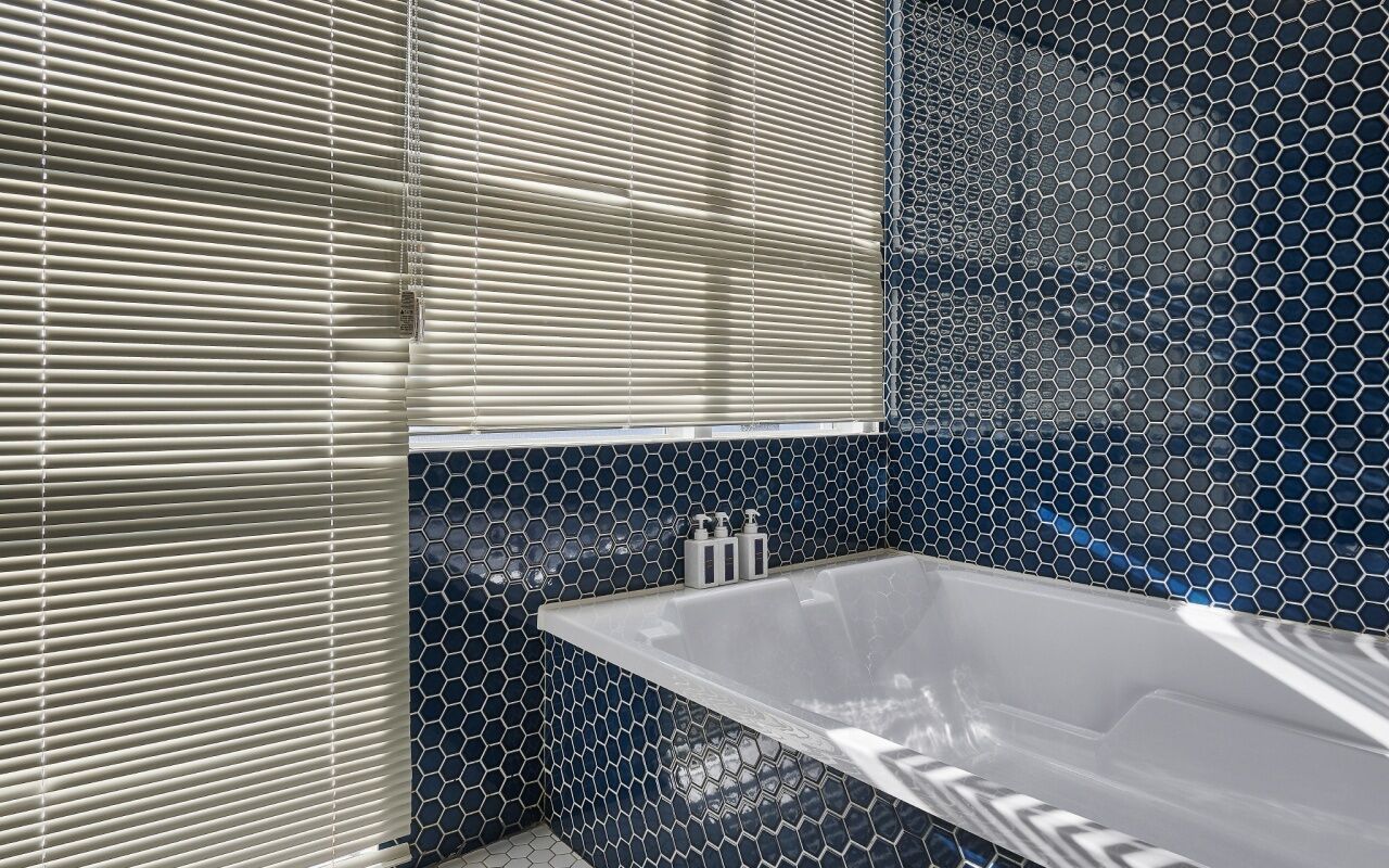 A bathroom with a bathtub, a window with blinds, and a tiled wall.