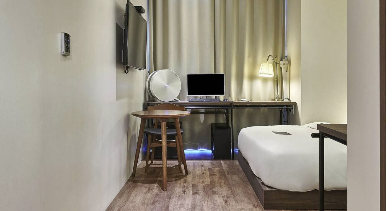 Habitación de hotel con cama, escritorio con computadora, silla, televisor y lámpara.