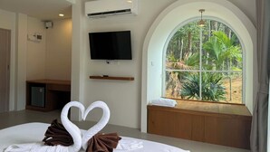 Habitación de hotel con cama, televisor, aire acondicionado, ventana con vista a vegetación y una decoración de toalla con forma de corazón.