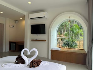 Ein Hotelzimmer mit Bett, Fernseher, Klimaanlage, einem Fenster mit Blick auf GrĂŒnflĂ€chen und einer Herz-förmigen Handtuchdekoration.