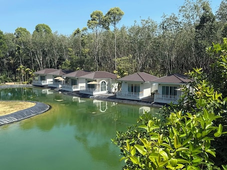 A row of white buildings with red roofs, a pond, and dense trees in the background.