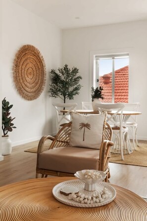 Ein Zimmer mit einem Korbstuhl, einem weißen Tisch mit einer dekorativen Platte, einer gewebten Wanddekoration und einer Topfpflanze.