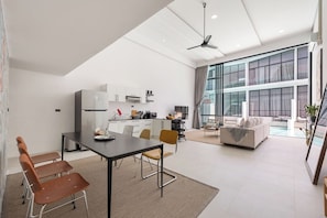 A modern kitchen with white cabinets, a dining area with a table and chairs, and a living area with a sofa and a view of the outdoors.