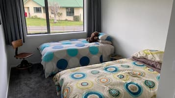Ein Zimmer mit zwei Betten, einem Fenster mit Vorhängen, einem kleinen Tisch und einem Stuhl.