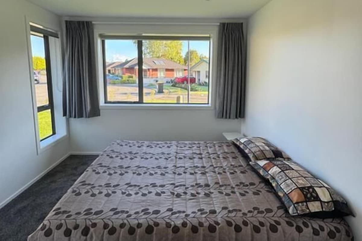 Quarto com cama grande, janela com cortinas e vista para uma área residencial.
